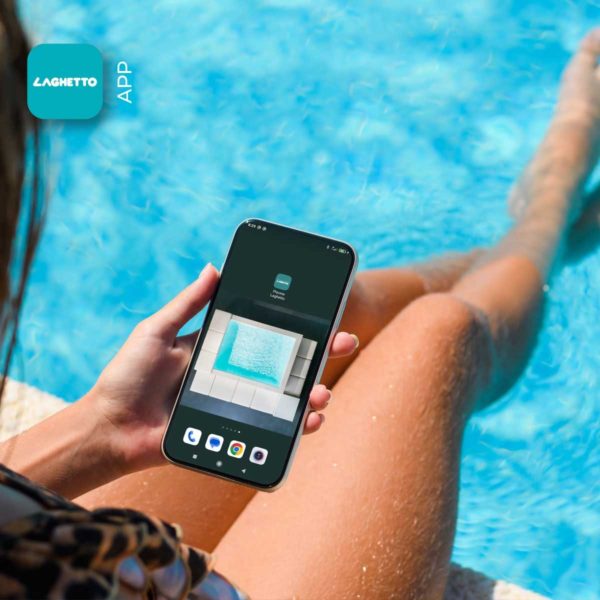 Una persona seduta sul bordo di una piscina, con le gambe immerse nell'acqua cristallina. La persona tiene in mano un telefono cellulare, mostrando uno screenshot di un'app con il logo "LAGHETTO APP". L'immagine trasmette una sensazione di relax e spensieratezza, tipica di una giornata trascorsa in piscina. L'attenzione è focalizzata sull'app "LAGHETTO APP", suggerendo che potrebbe essere utile per gestire o controllare la piscina.