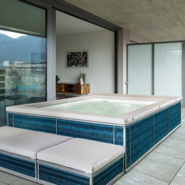 Una piscina Laghetto Ninfea Thiny idromassaggio rettangolare, con rivestimento esterno in corda colore Deep Blue intrecciato, è posizionata in una terrazza coperta. L'acqua della piscina è azzurro chiaro e sembra invitante. L'ambiente è elegante e raffinato, con elementi di design moderno.