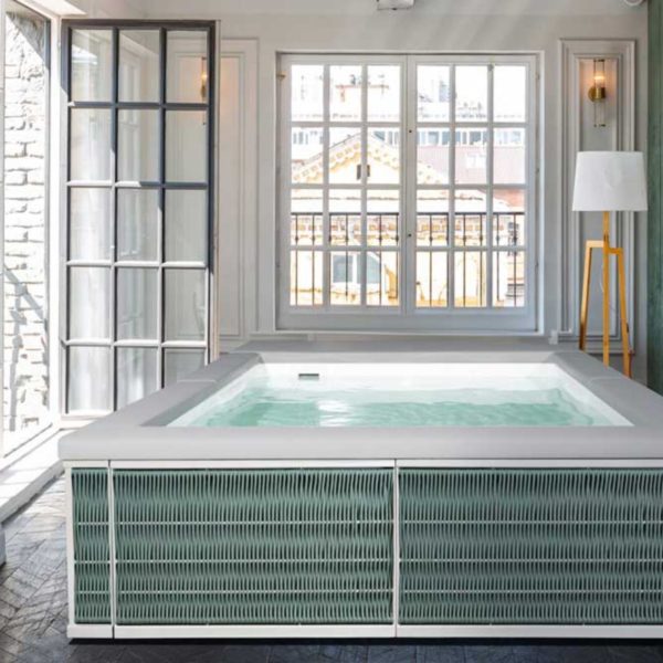 Una piscina Laghetto Ninfea Thiny idromassaggio quadrata, con rivestimento esterno in corda colore Olive Green intrecciato, è posizionata in un ambiente interno luminoso con grandi finestre. L'acqua della piscina è azzurro chiaro e sembra invitante. L'ambiente è elegante e raffinato, con elementi di design moderno e classico.