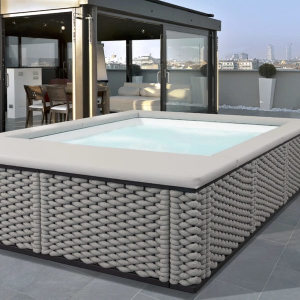 Una piscina Laghetto Ninfea idromassaggio quadrata, con rivestimento esterno in corda colore SUNRISE intrecciato, è posizionata in una terrazza scoperta. L'acqua della piscina è azzurro chiaro e sembra invitante. L'ambiente è elegante e raffinato, con elementi di design moderno.