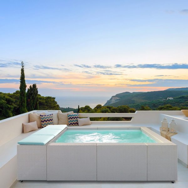 Una piscina fuori terra Laghetto Playa con idromassaggio integrato è posizionata su una terrazza panoramica con vista sul mare e sulle montagne. La piscina ha un rivestimento esterno in vimini sintetico chiaro e cuscini di seduta coordinati. L'acqua è di un colore azzurro chiaro e sembra invitante. Il cielo è sereno con un tramonto all'orizzonte. L'atmosfera è rilassante e lussuosa.