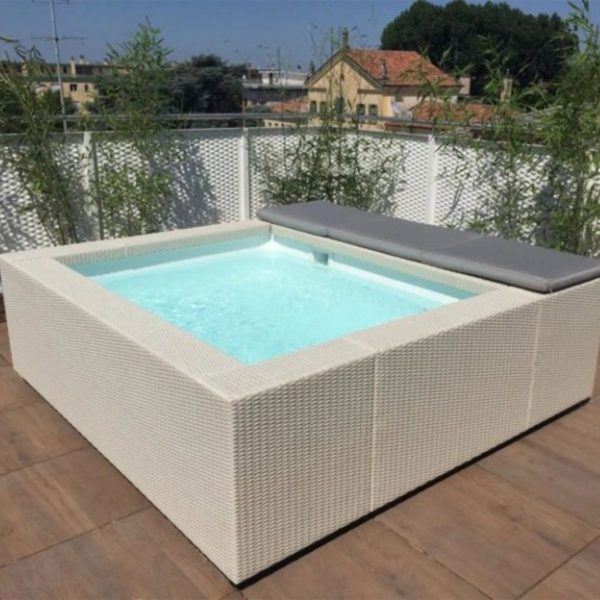 Una piscina fuori terra modello Laghetto Playa modello Playa 1 (220x220 cm) di forma quadrata con idromassaggio integrato è posizionata su una terrazza con pavimentazione in piastrelle. La piscina ha un rivestimento esterno in vimini sintetico chiaro e un cuscino di seduta grigio su un lato. L'acqua è di un colore azzurro chiaro. Sullo sfondo, si vedono edifici residenziali e vegetazione.